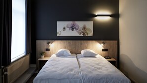 Une chambre d’hôtel avec deux lits, un mur de couleur sombre, une fenêtre avec des rideaux, un luminaire fixé au mur et une œuvre d’art florale au-dessus des lits.