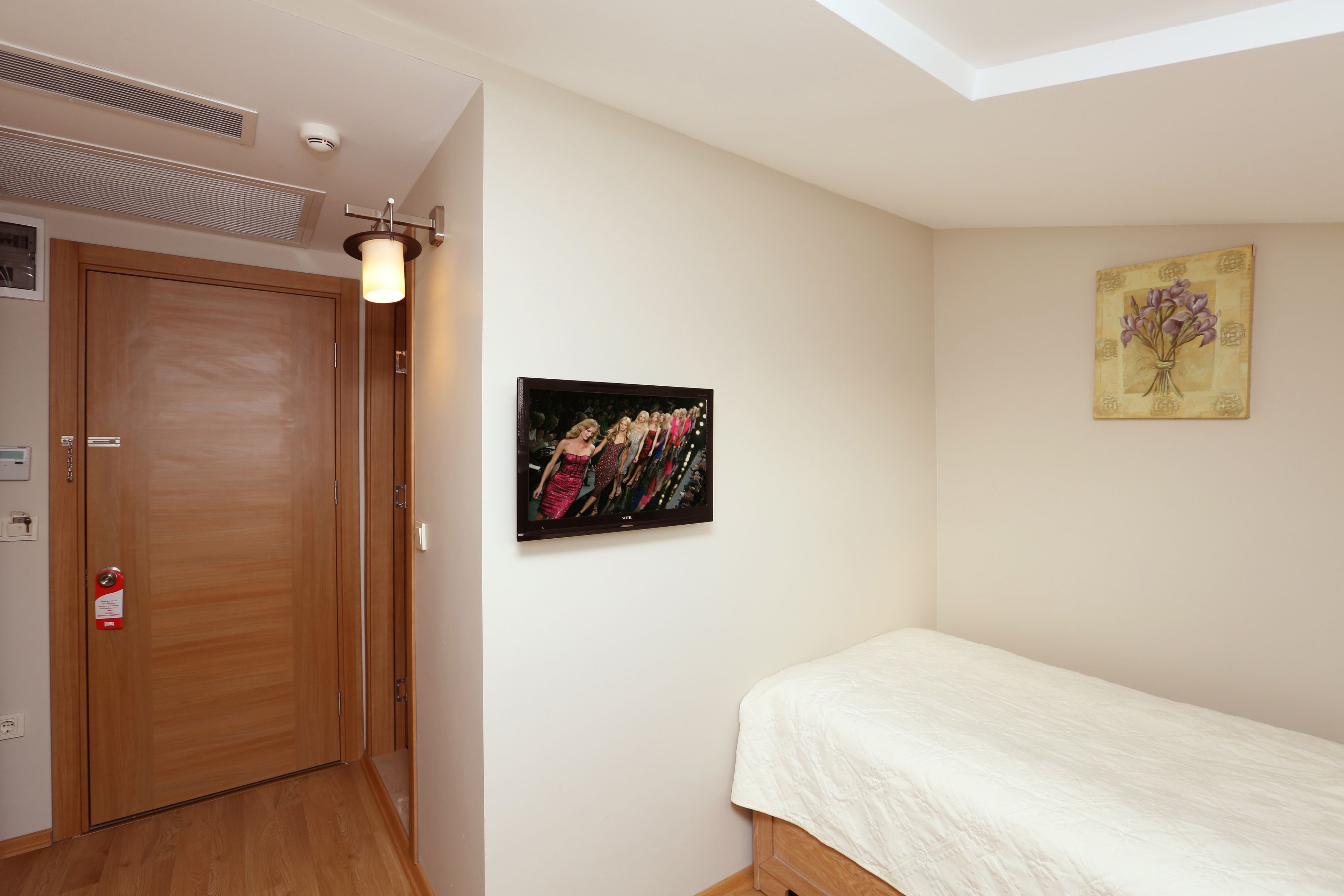 Ein Schlafzimmer mit Holztür, einem Bett mit weißer Bettdecke, einem an der Wand hängenden Bild und einer Deckenleuchte.