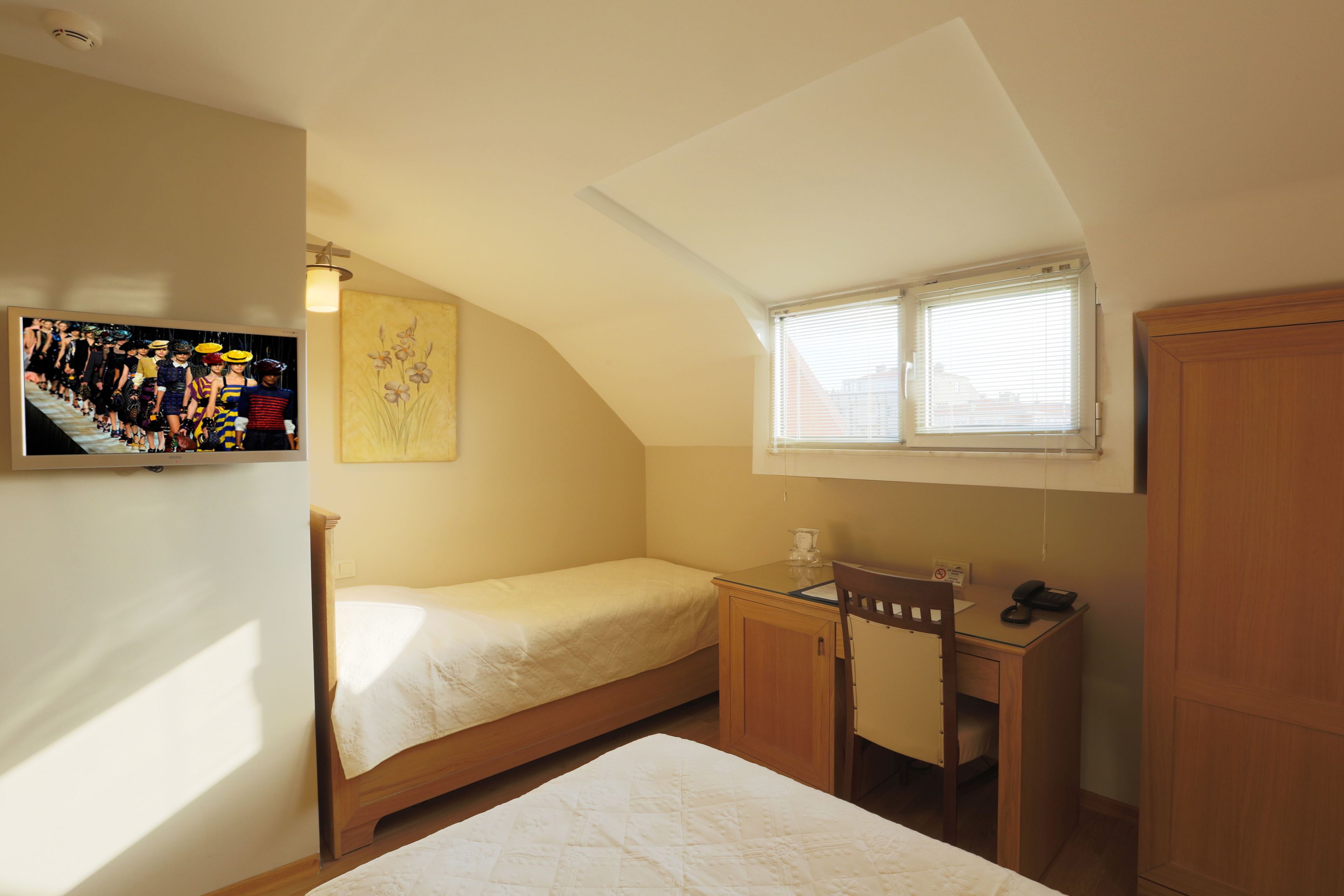 A small, well-lit bedroom with a single bed, a desk with a chair, a window with blinds, and a wall-mounted TV displaying a children's show.