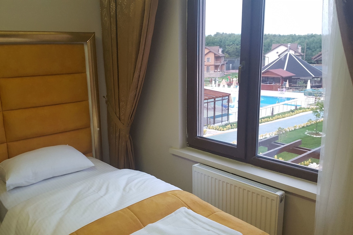 Quarto de hotel com cama, mesa de cabeceira e janela com vista para uma piscina e edifícios.