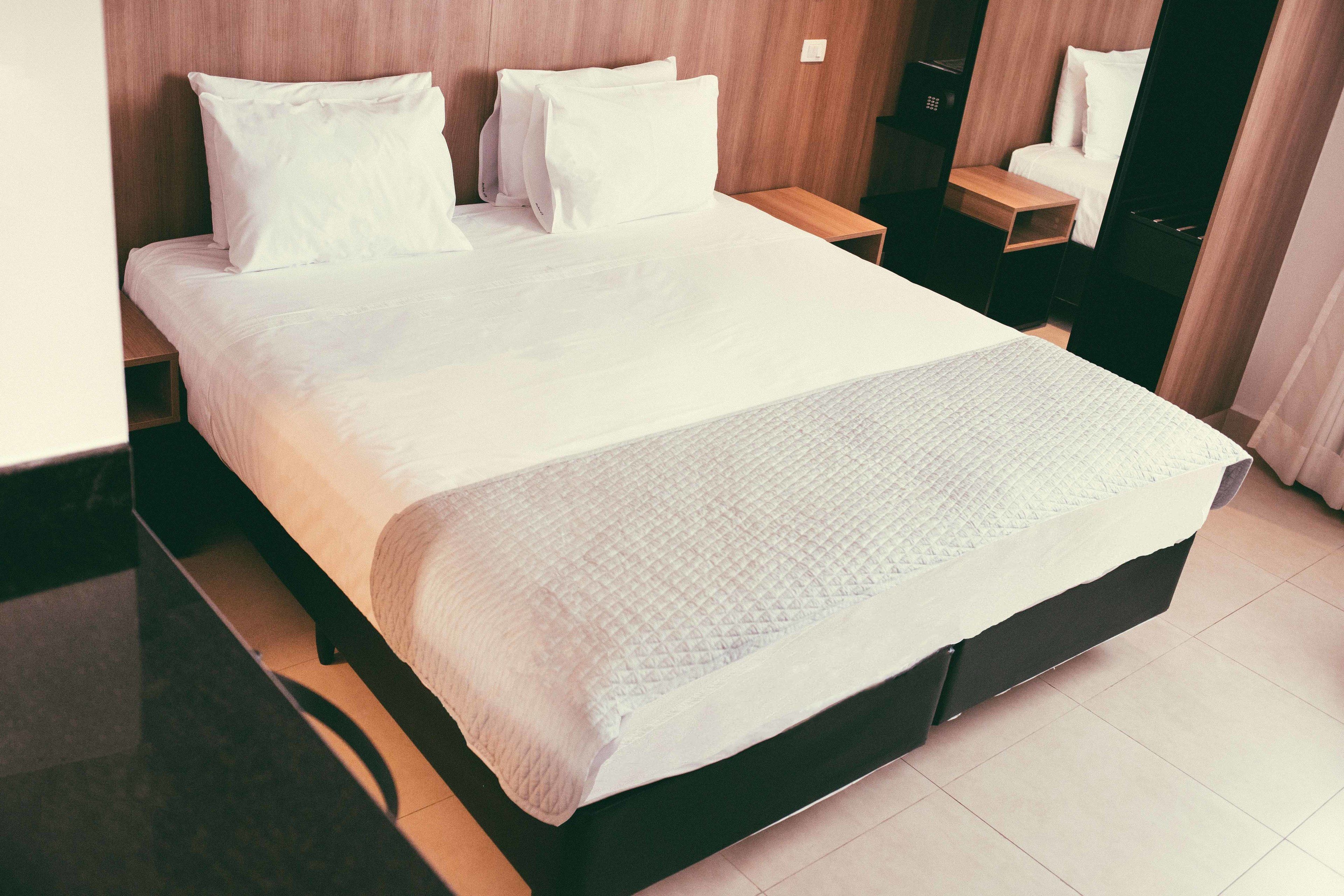 Una cama bien hecha con cubrecama acolchado y almohadas, cabecera de madera, mesita de noche y velador con lámpara.