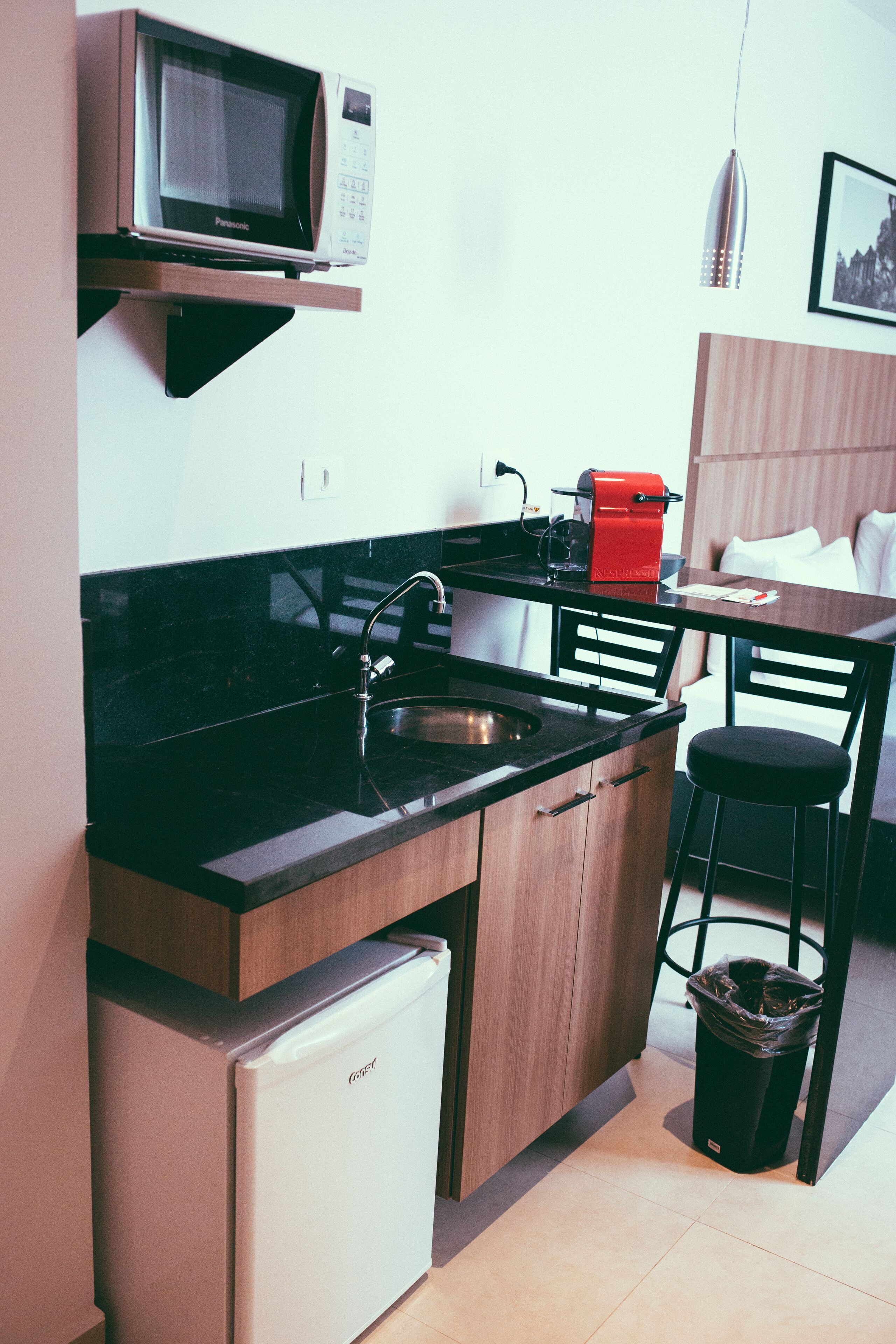 Cozinha compacta com micro-ondas, geladeira e pia. Há também uma pequena lixeira e uma cadeira.