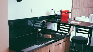 Cozinha compacta com micro-ondas, geladeira e pia. Há também uma pequena lixeira e uma cadeira.