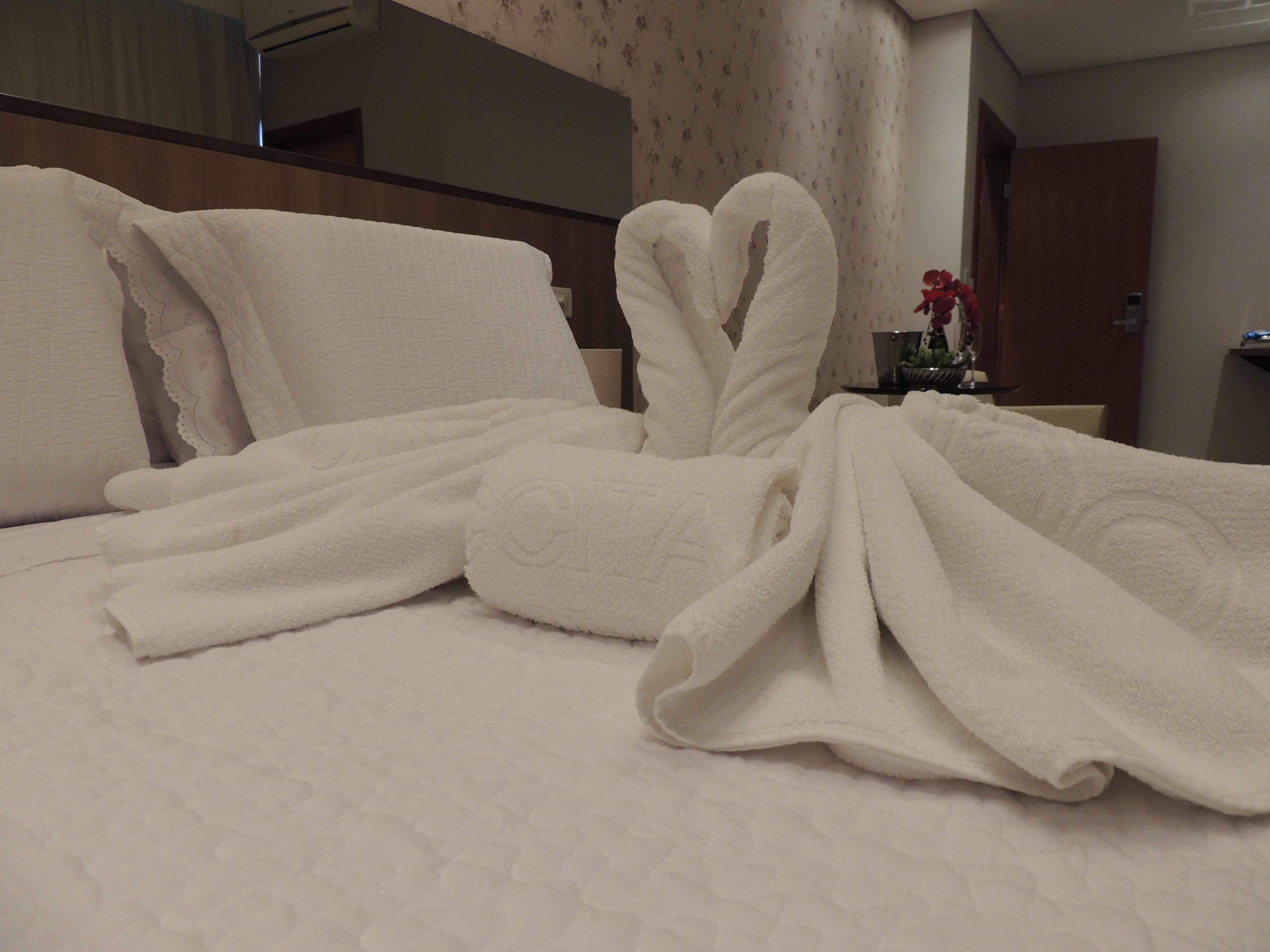 A neatly made bed with a white quilt and two towels folded in a heart shape.