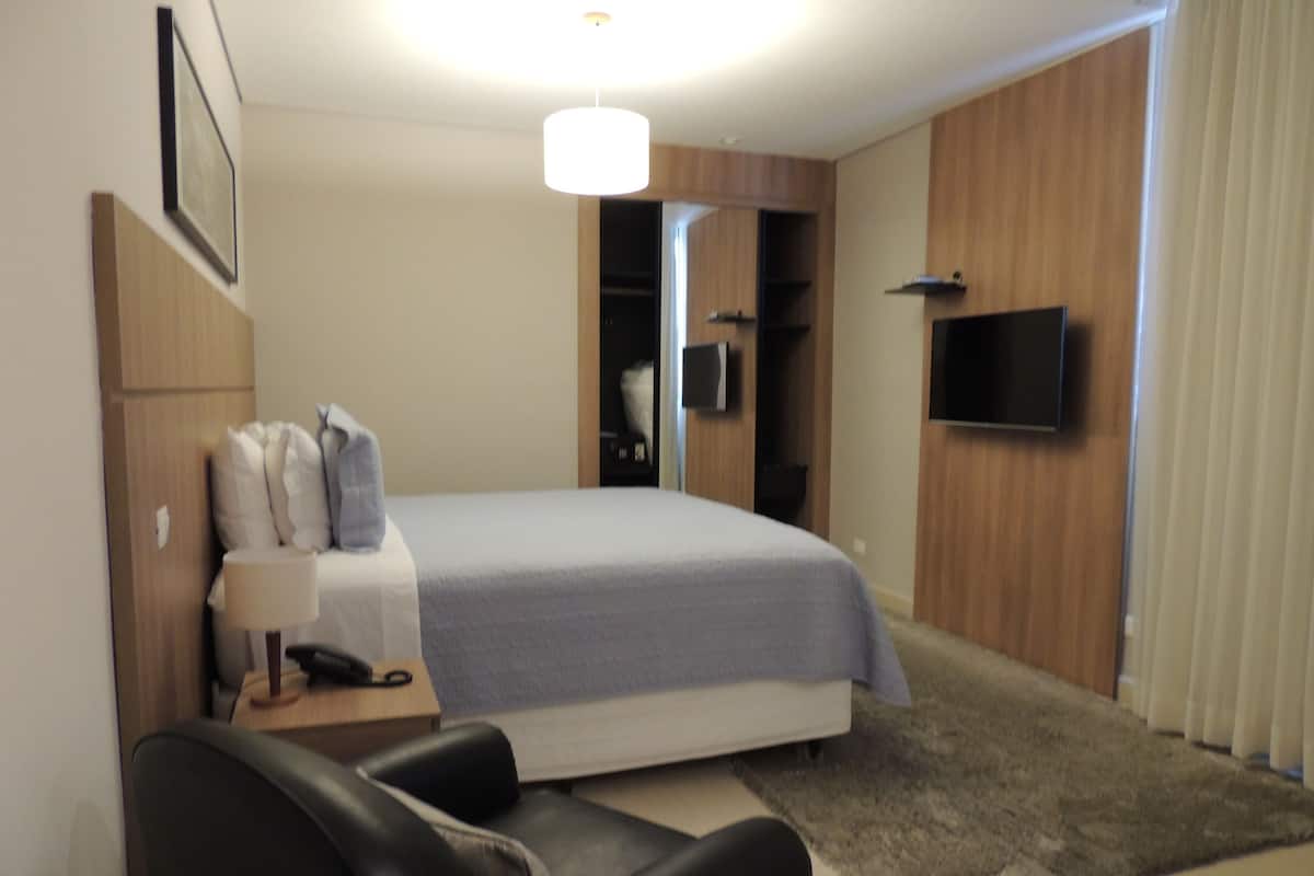 A modern hotel room with a bed, a desk with a lamp, a chair, a TV, and a wardrobe.
