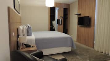 A modern hotel room with a bed, a desk with a lamp, a chair, a TV, and a wardrobe.