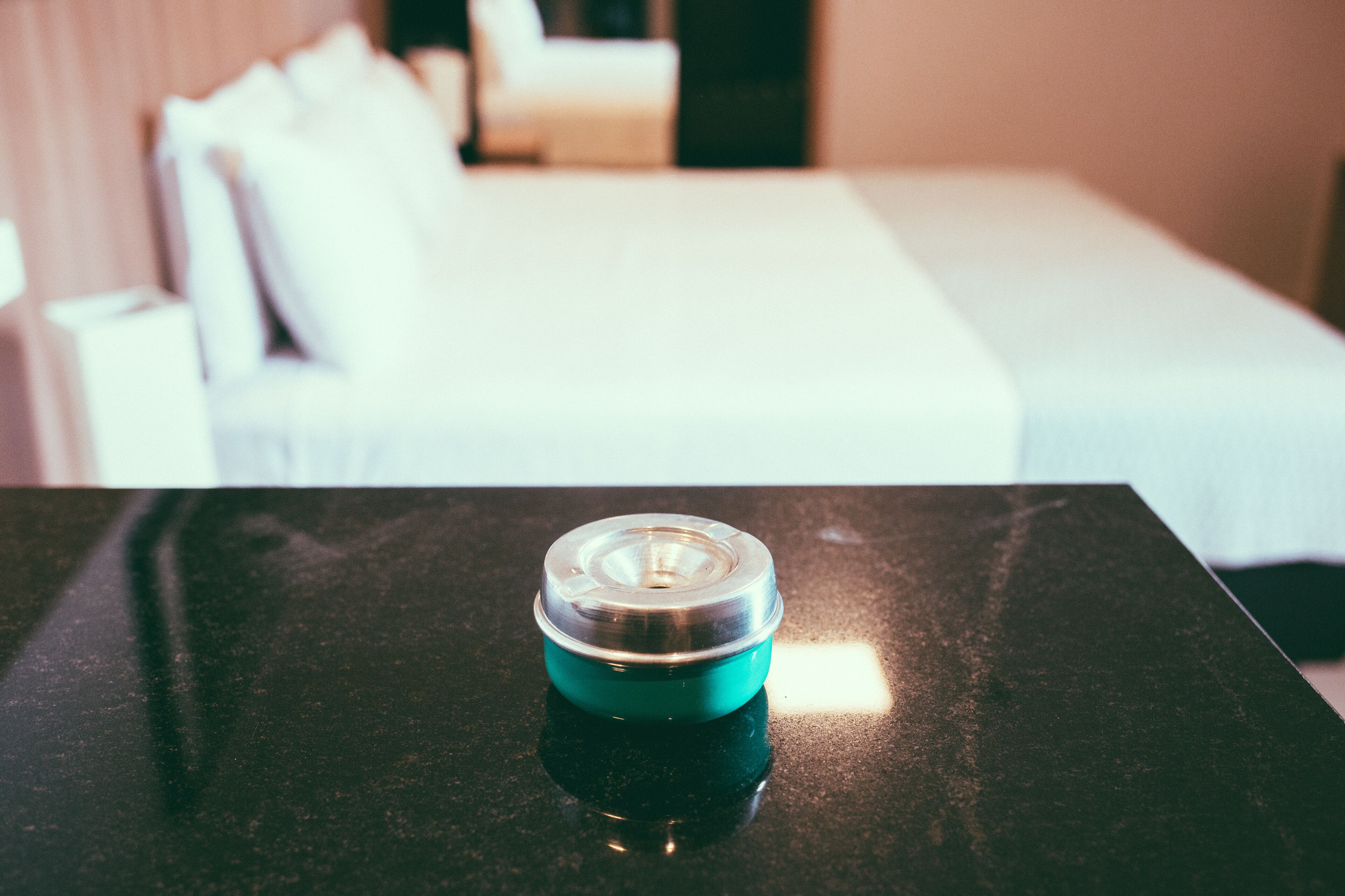 A small, teal-colored container on a black surface, likely a table, with a blurred background suggesting a hotel room with a bed and pillows.