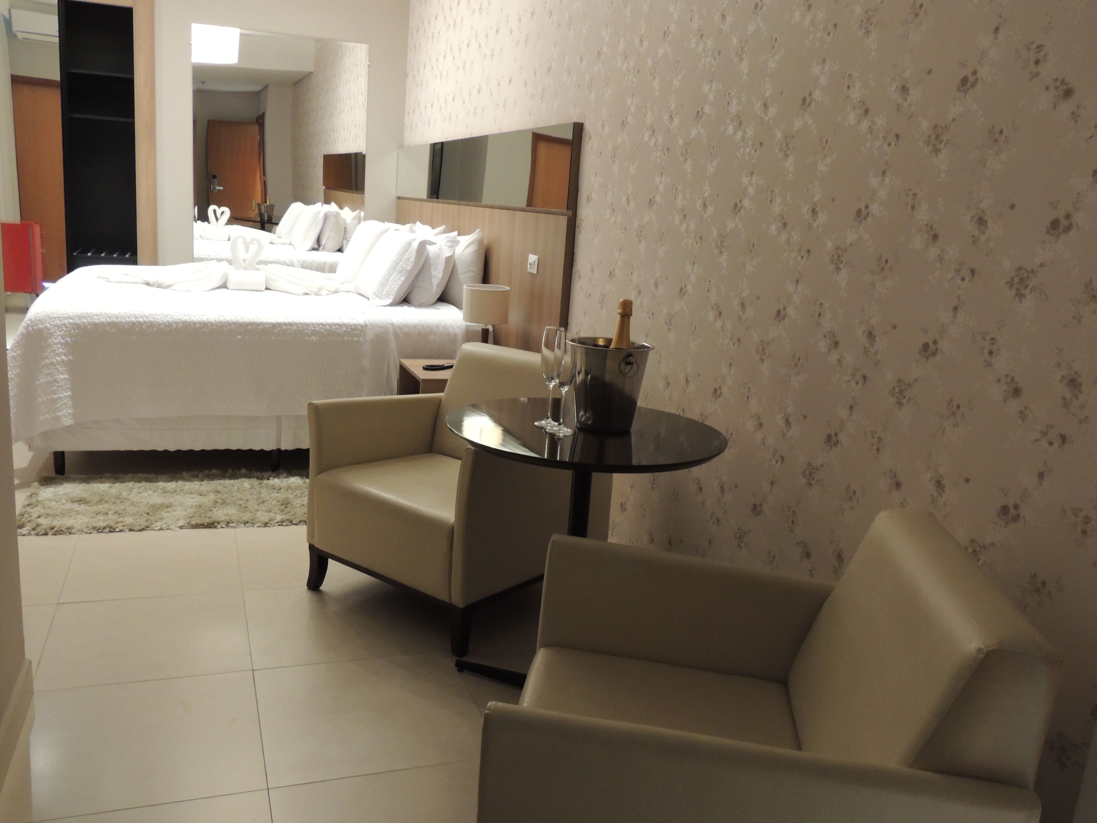 A hotel room with a bed, two armchairs, a small table with a bottle of champagne, and a mirror on the wall.