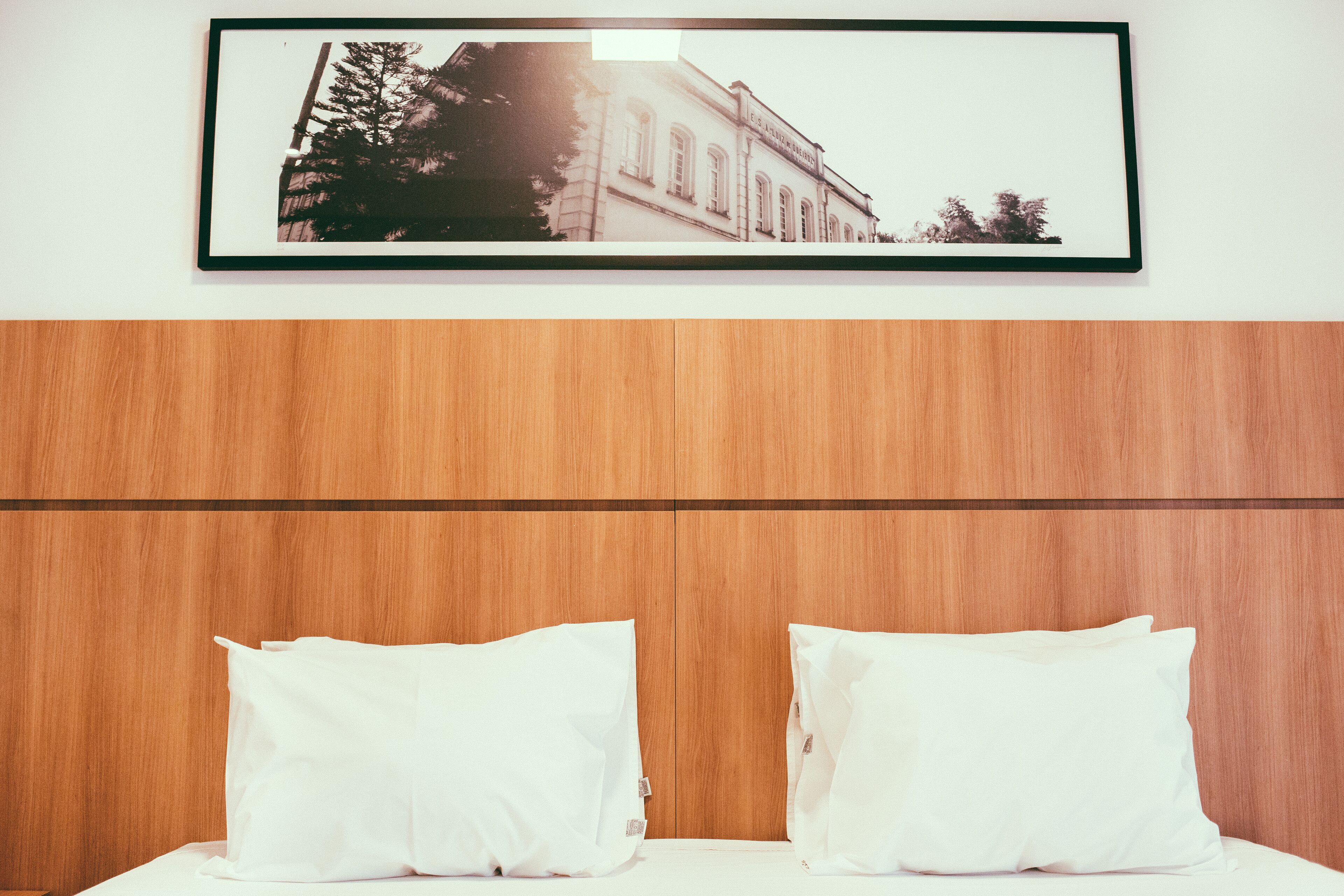 A hotel room with a wooden headboard, two white pillows, and a framed picture of a building on the wall.