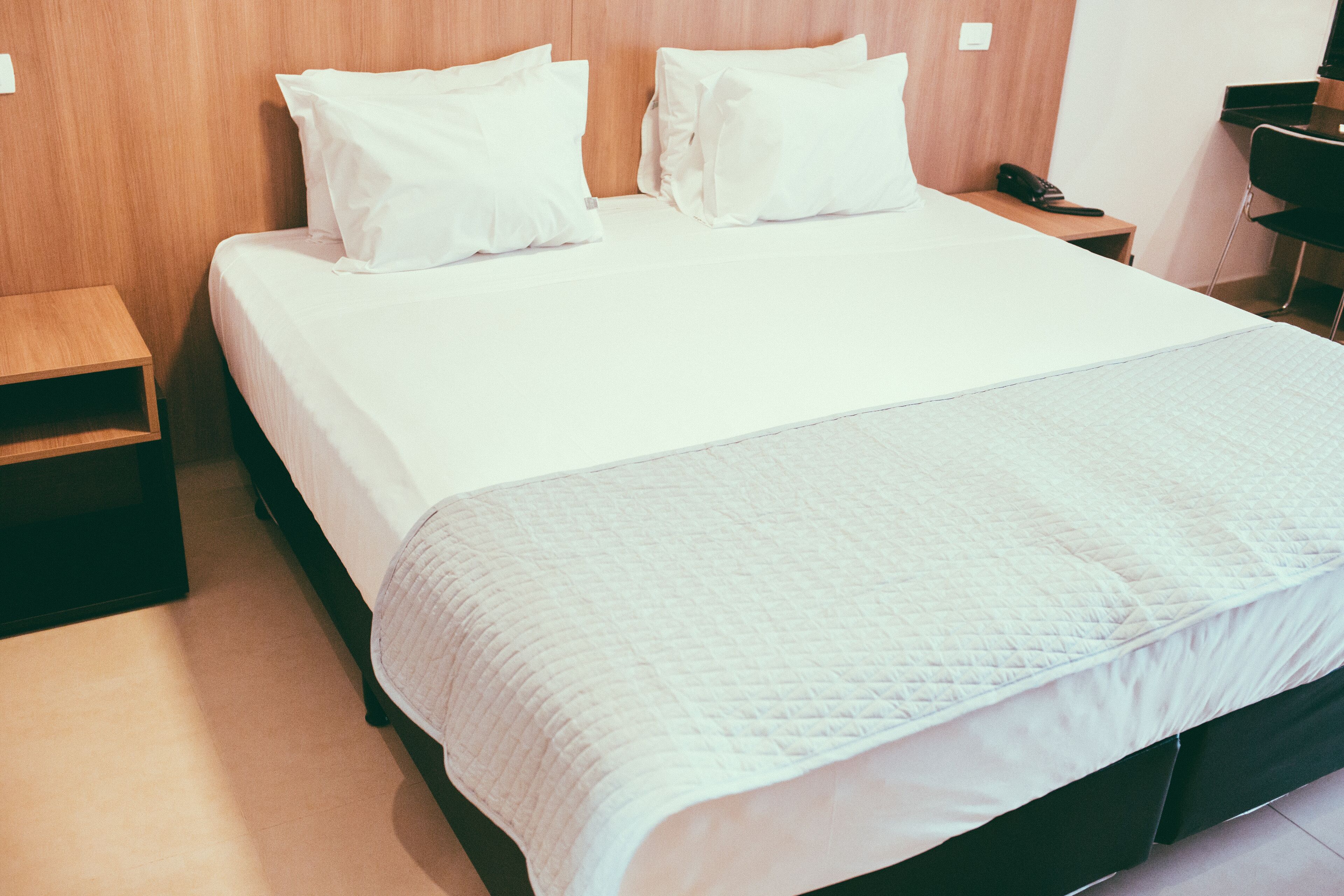 Una cama bien hecha con sábanas y almohadas blancas, un cabecero de madera, una mesita de noche de madera y una silla negra.
