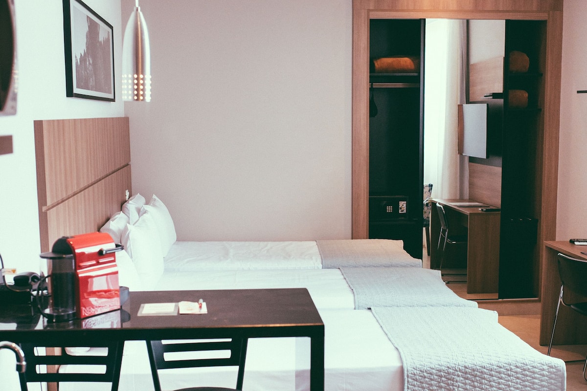 A modern hotel room with a bed, a desk, and a chair. There is a coffee maker on the desk and a trash can next to it.