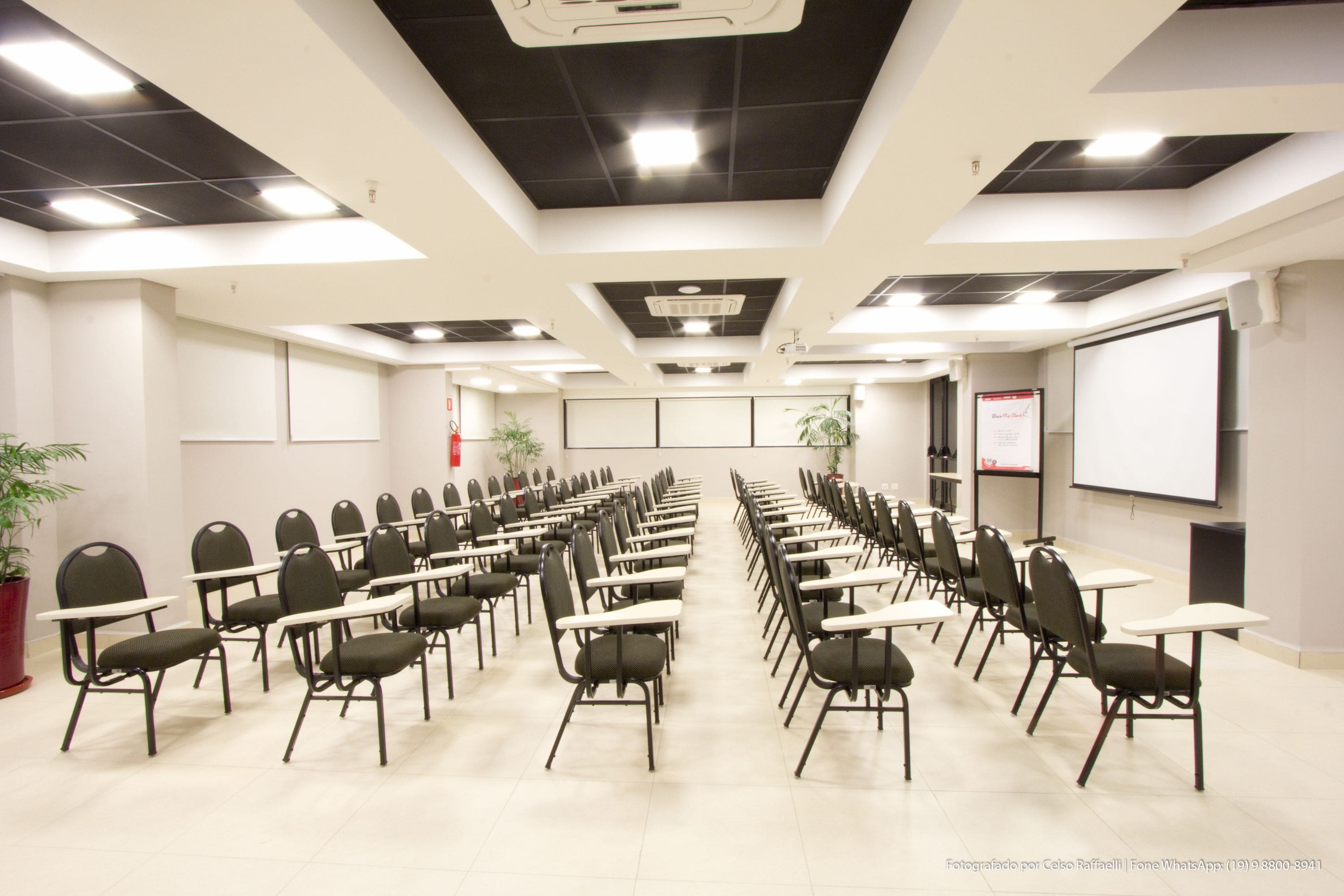 Uma sala de conferência com fileiras de cadeiras e mesas, uma tela de projetor e luminárias no teto.