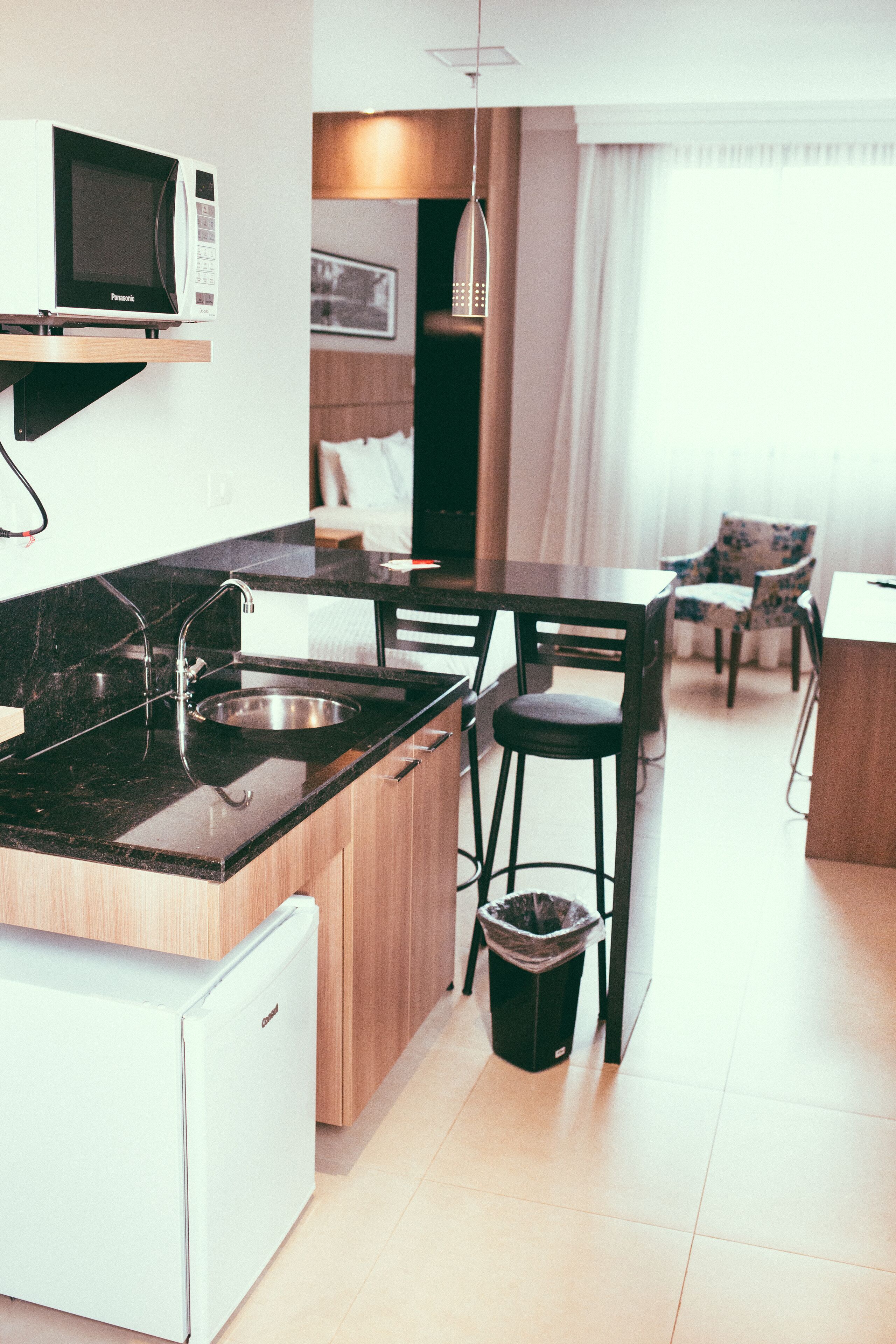 A compact kitchen with a microwave, sink, and small refrigerator. There is a dining area with a table and chairs, and a trash bin.