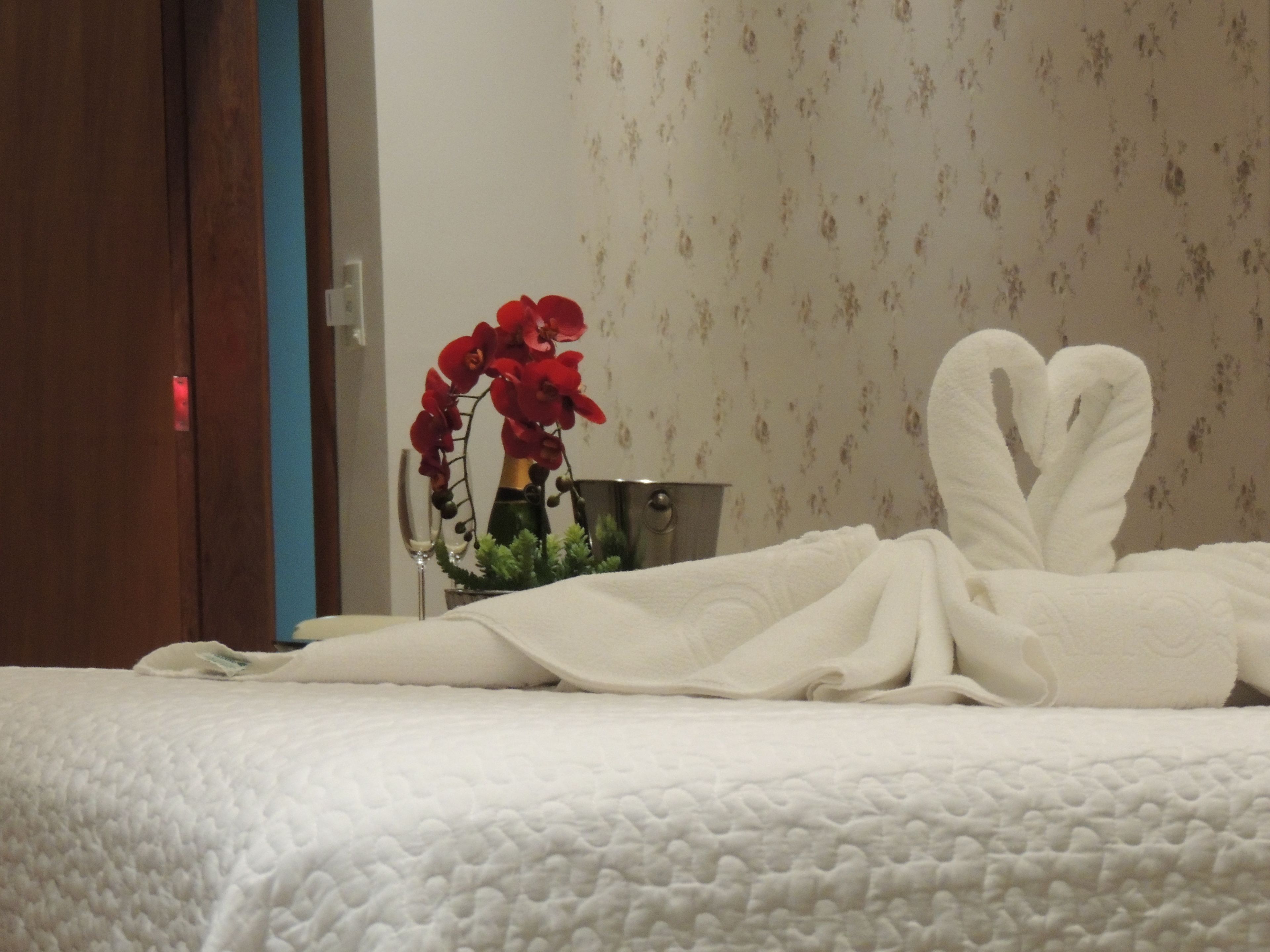 A neatly made bed with white towels arranged in a heart shape, a vase with red flowers, and a metal bucket on a bedside table.