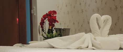 Un lit bien fait, avec des serviettes blanches disposées en forme de cœur, un vase rempli de fleurs rouges et un seau en métal posé sur une table de chevet.