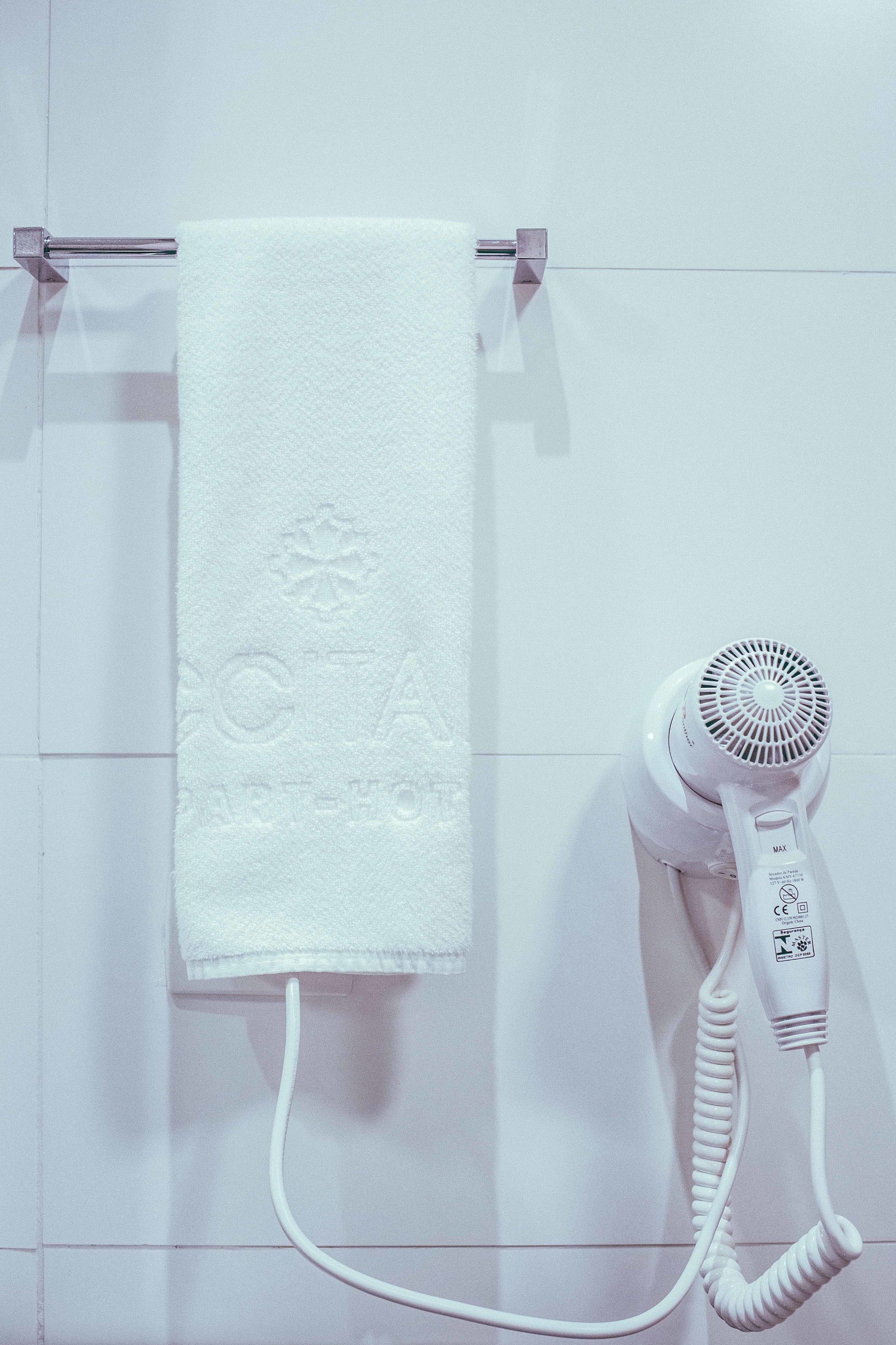 A white towel hanging on a metal towel rack, a white hairdryer with a cord, and a white wall with a subtle embossed design.
