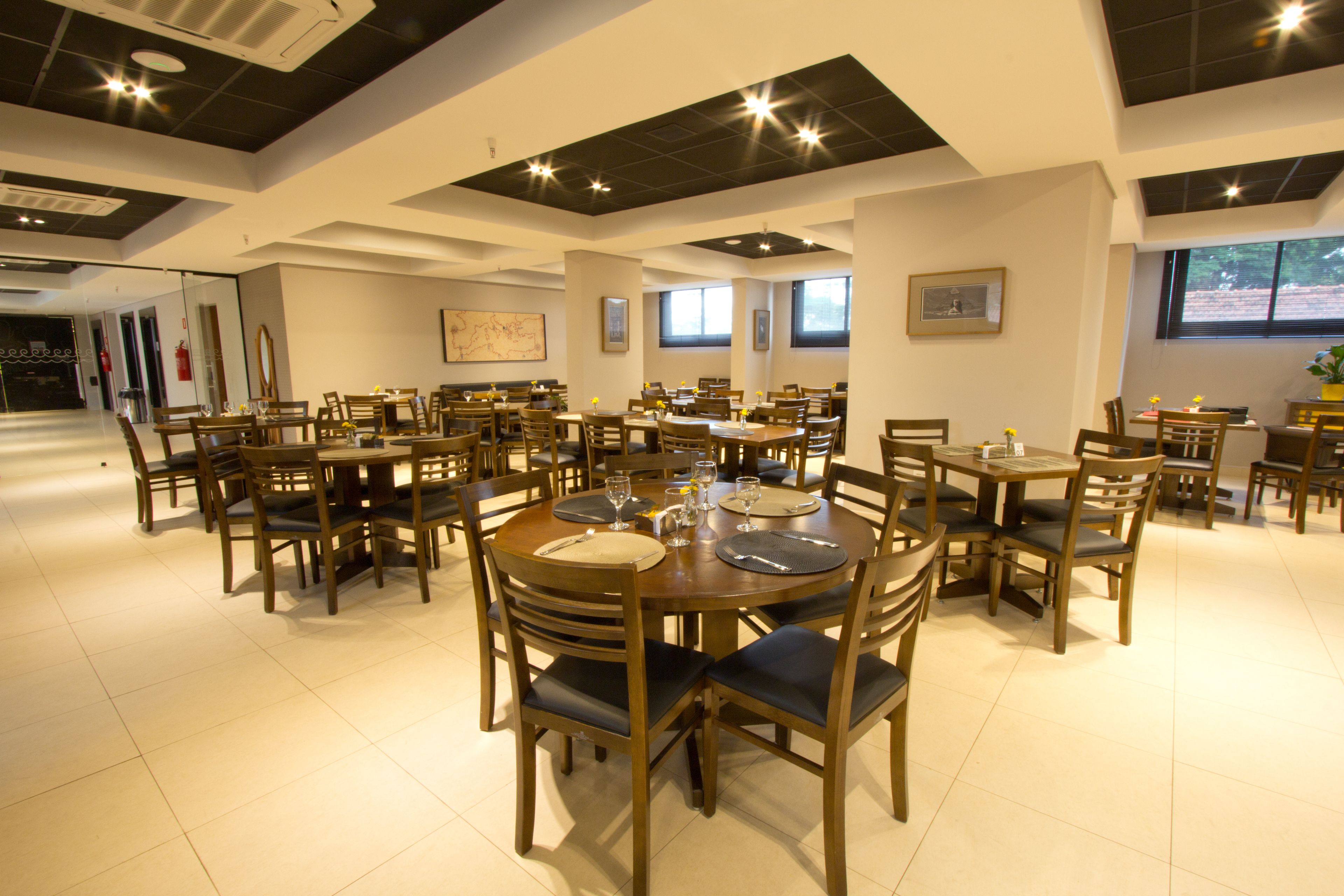 A spacious dining area with multiple round tables and chairs, featuring a modern ceiling design and large windows allowing natural light.