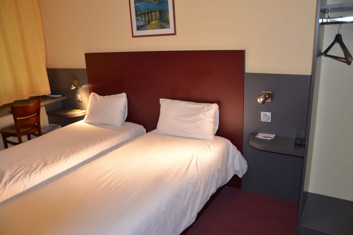 A hotel room with two single beds, a wooden chair, a nightstand, and a small table with a lamp.
