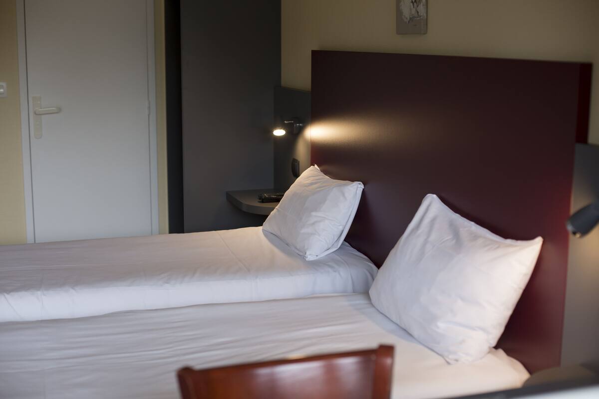 A neatly made bed with white linens and two pillows, a wooden chair in front, and a nightstand with a lamp.