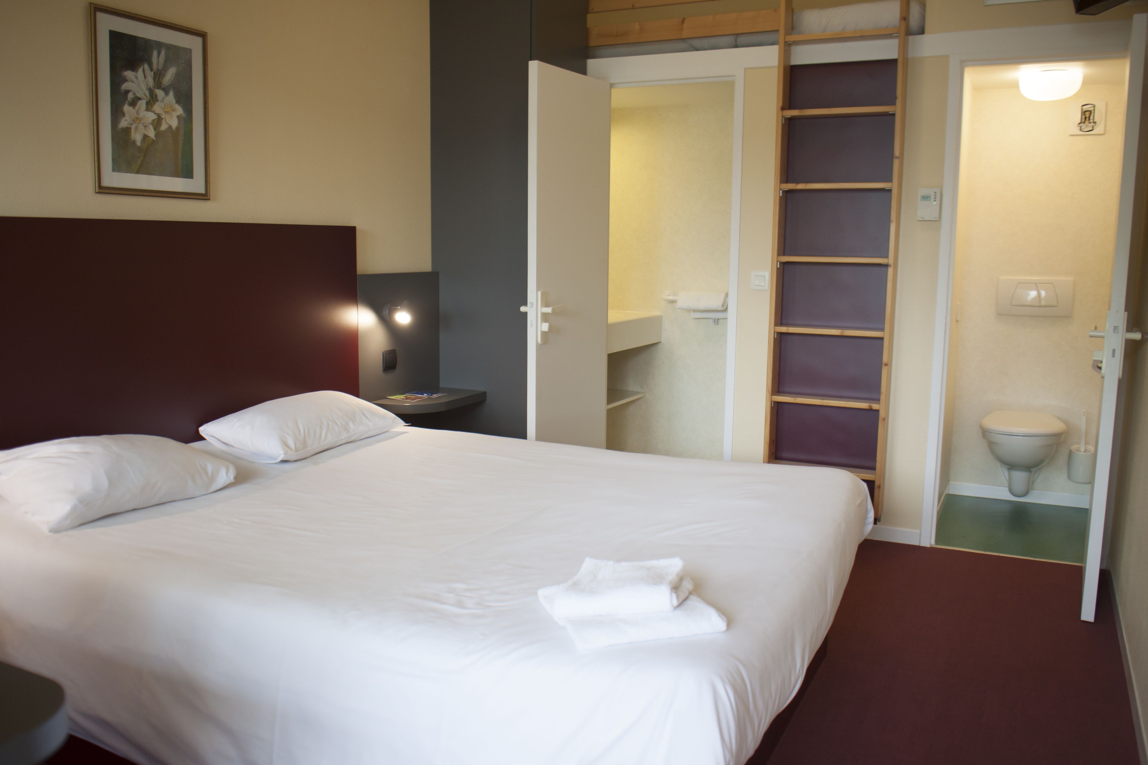 A neatly made bed with white linens and pillows, a bedside table with a lamp, and an open closet revealing shelves and a ladder.