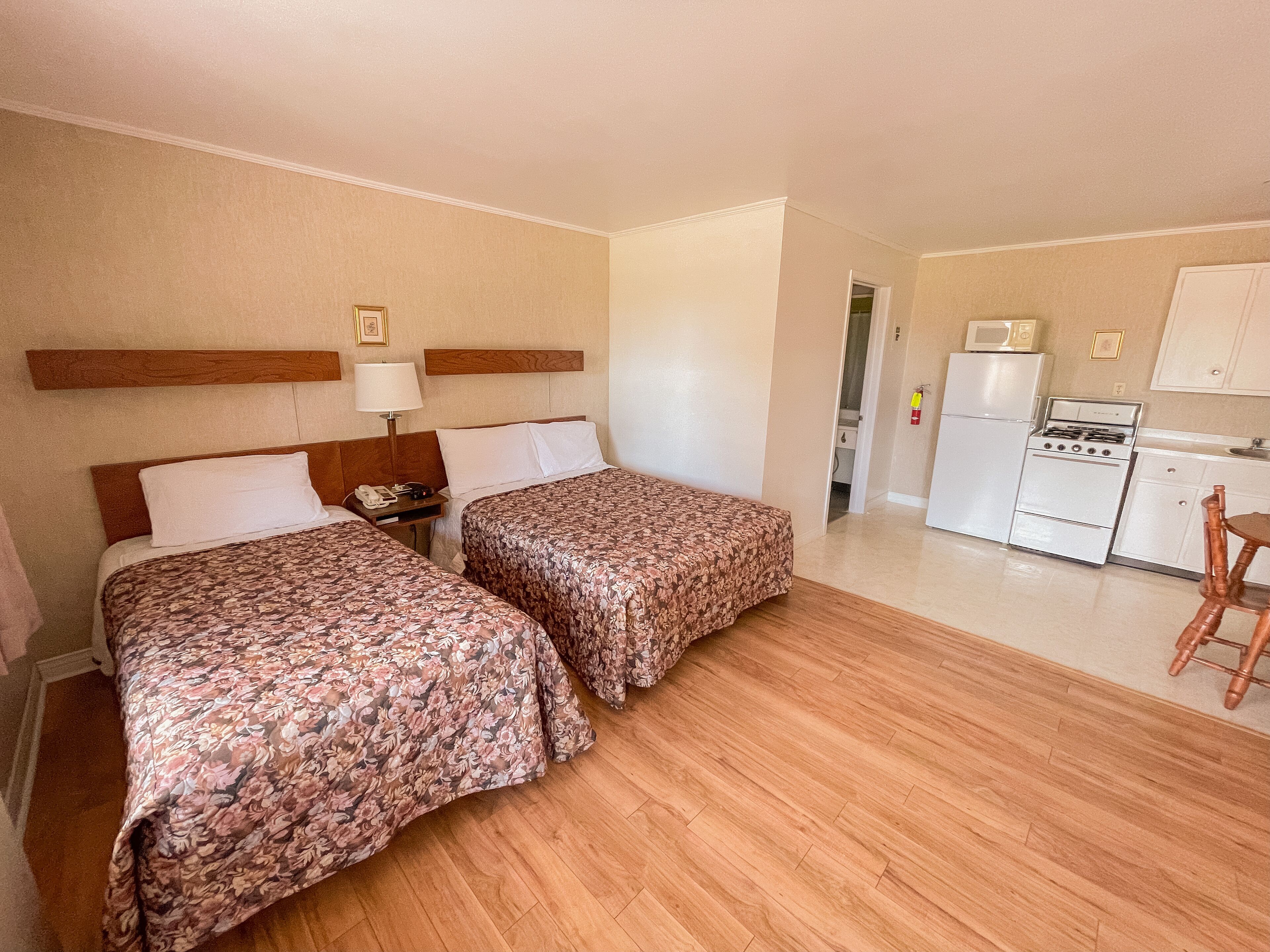 Une chambre d’hôtel avec deux lits, une tête de lit en bois, une table de chevet avec une lampe, un petit bureau et une kitchenette équipée d ’une cuisinière et d’un réfrigérateur.