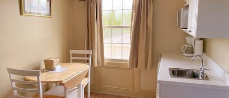 Una pequeña cocina con ventana, una mesa con sillas, un refrigerador blanco y una cafetera.
