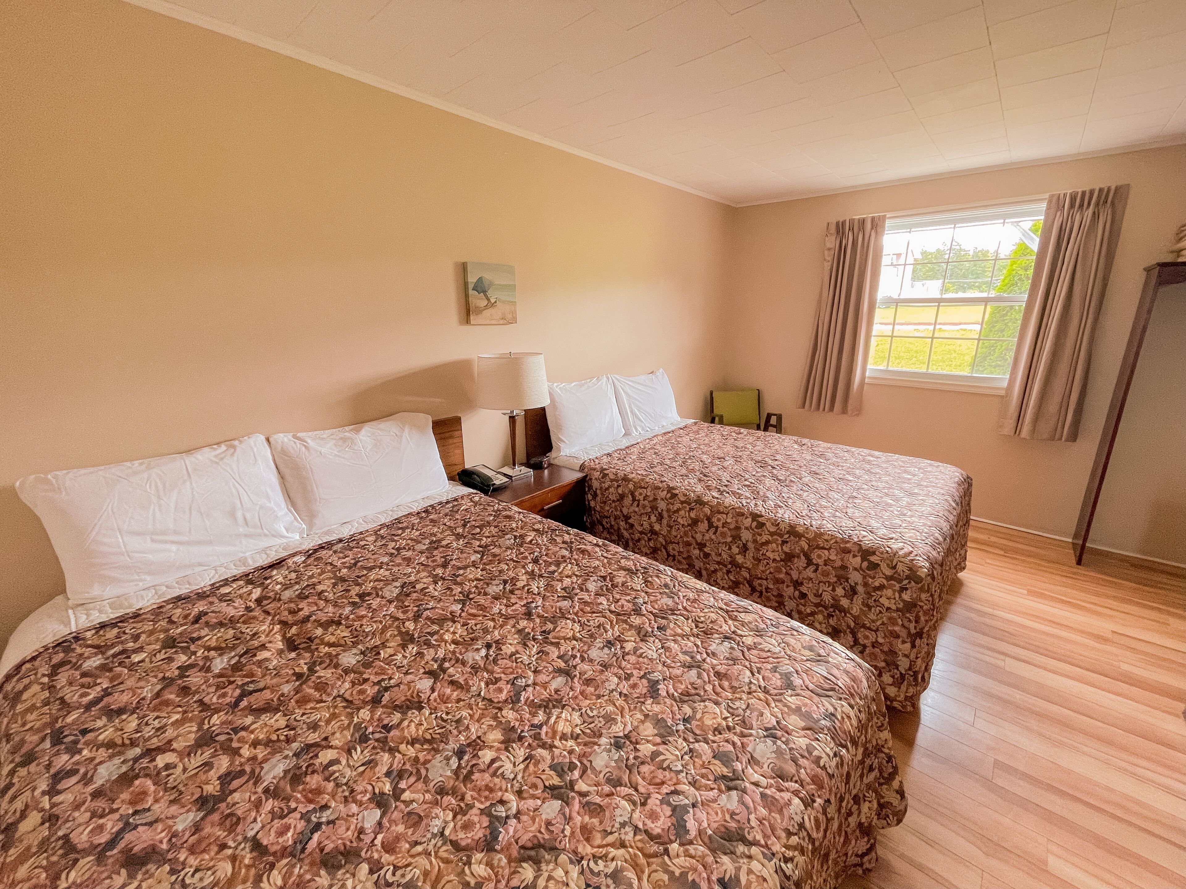 A hotel room with two beds, a nightstand, a lamp, a chair, a window with curtains, and a painting on the wall.