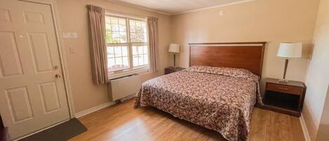 Une chambre avec un plancher en bois, un lit recouvert d’une couvre-lit à motifs floraux, une fenêtre avec des rideaux et deux tables de chevet avec des lampes.