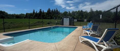 A swimming pool with lounge chairs, a small shed, and a fenced area with trees in the background.