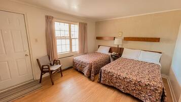 A room with two beds, a chair, a window with curtains, a bedside table with a lamp, and a door.