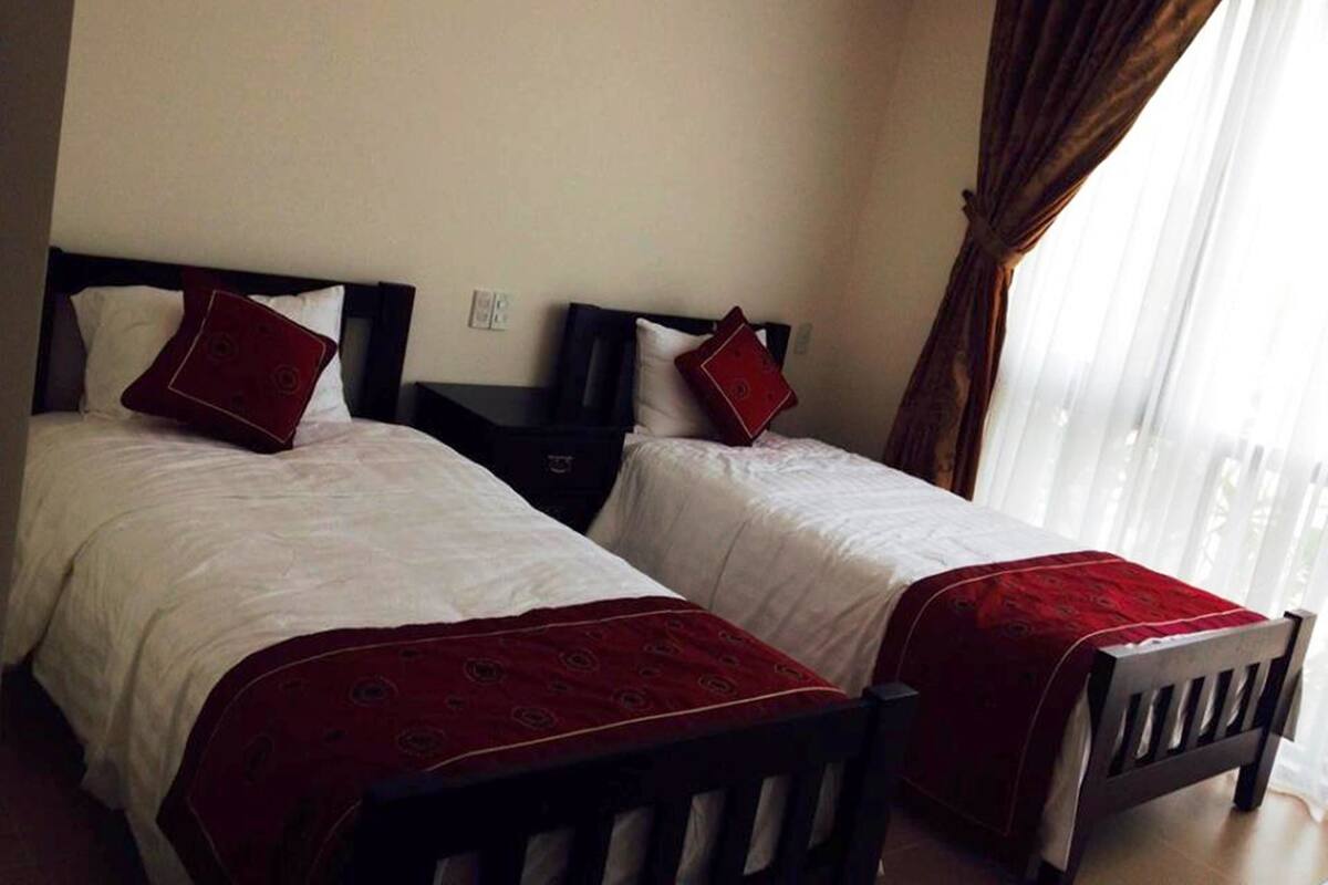 A hotel room with two single beds, each with a red bedspread and matching pillows. There is a wooden nightstand between the beds, and a window with curtains to the right.