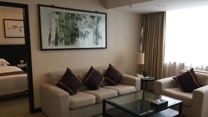 Habitación de hotel con sofá beige, una mesita de centro de vidrio, una obra de arte enmarcada en la pared y una cama con cabecera.