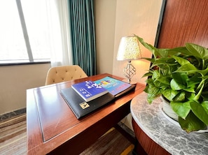 Habitación de hotel con escritorio de madera, una computadora portátil, lámpara, planta en maceta y ventana con cortinas.