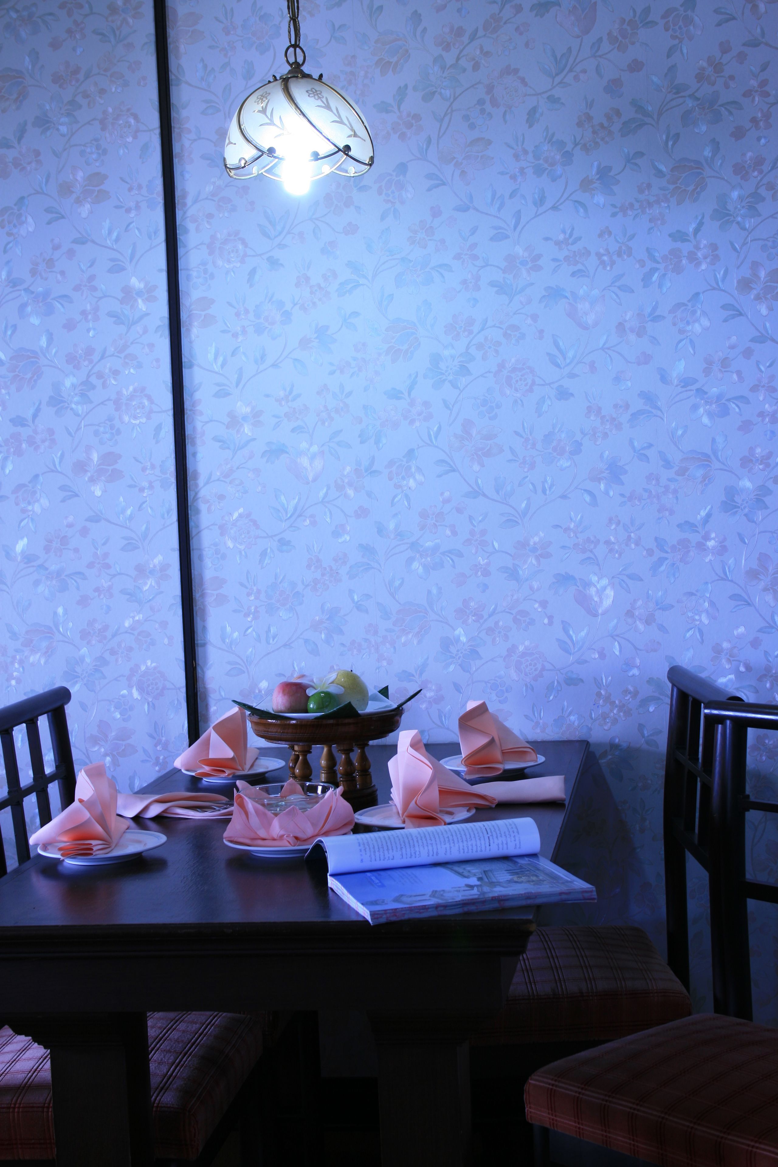 A dining table with a floral wallpaper background, a hanging light fixture, and neatly arranged plates, napkins, and a book.
