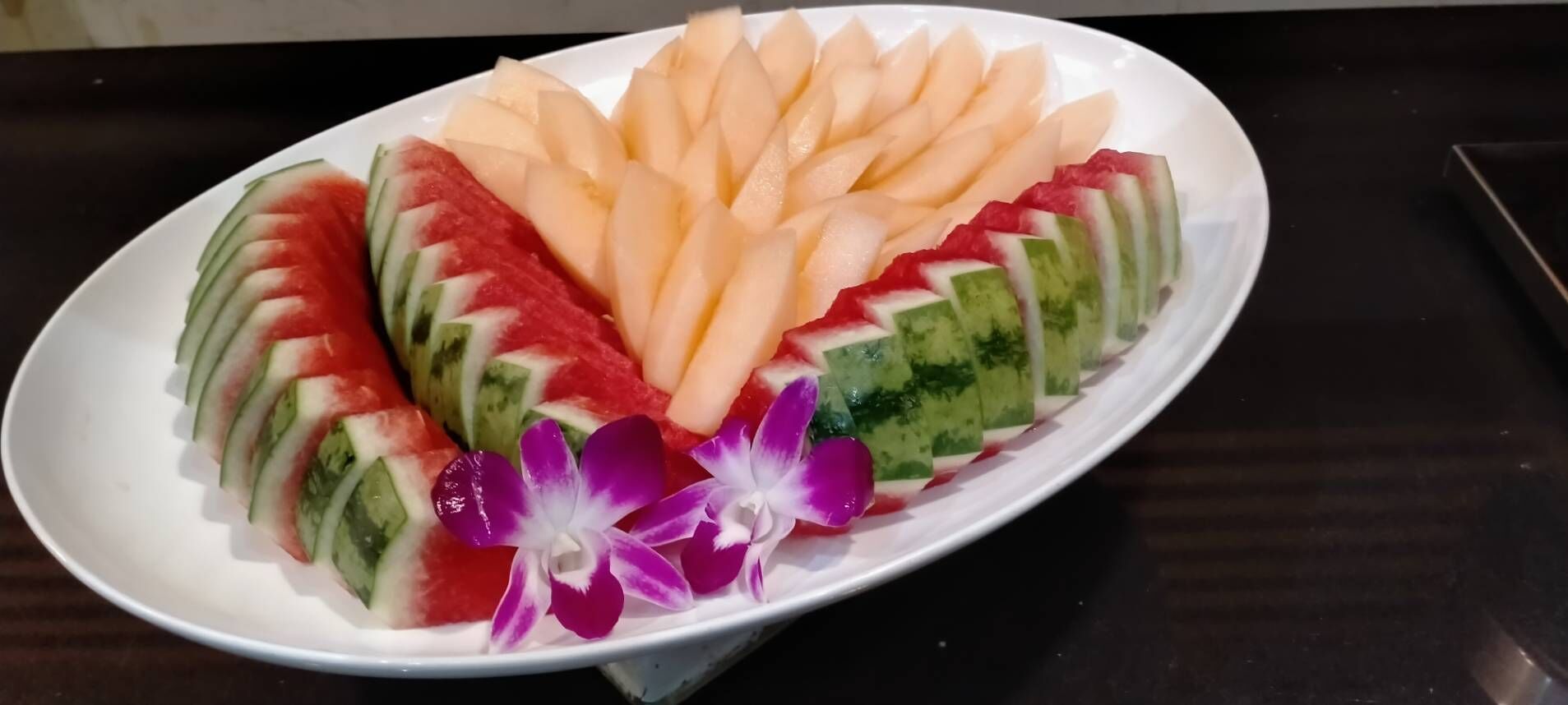 Una fuente con rebanadas de sandía y melón dispuestas en forma de abanico, adornada con orquídeas moradas.