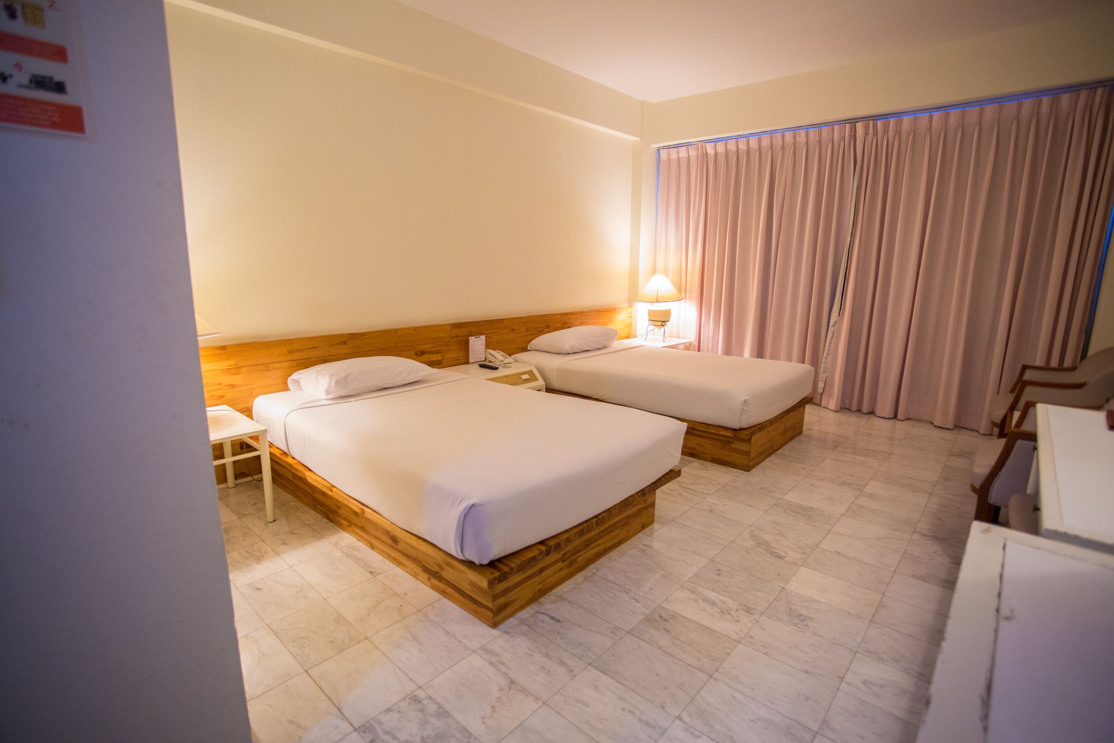 A hotel room with two wooden beds, a small table, a lamp, and a curtain.