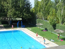 Una piscina con solárium de baldosas, rodeada de césped y árboles.