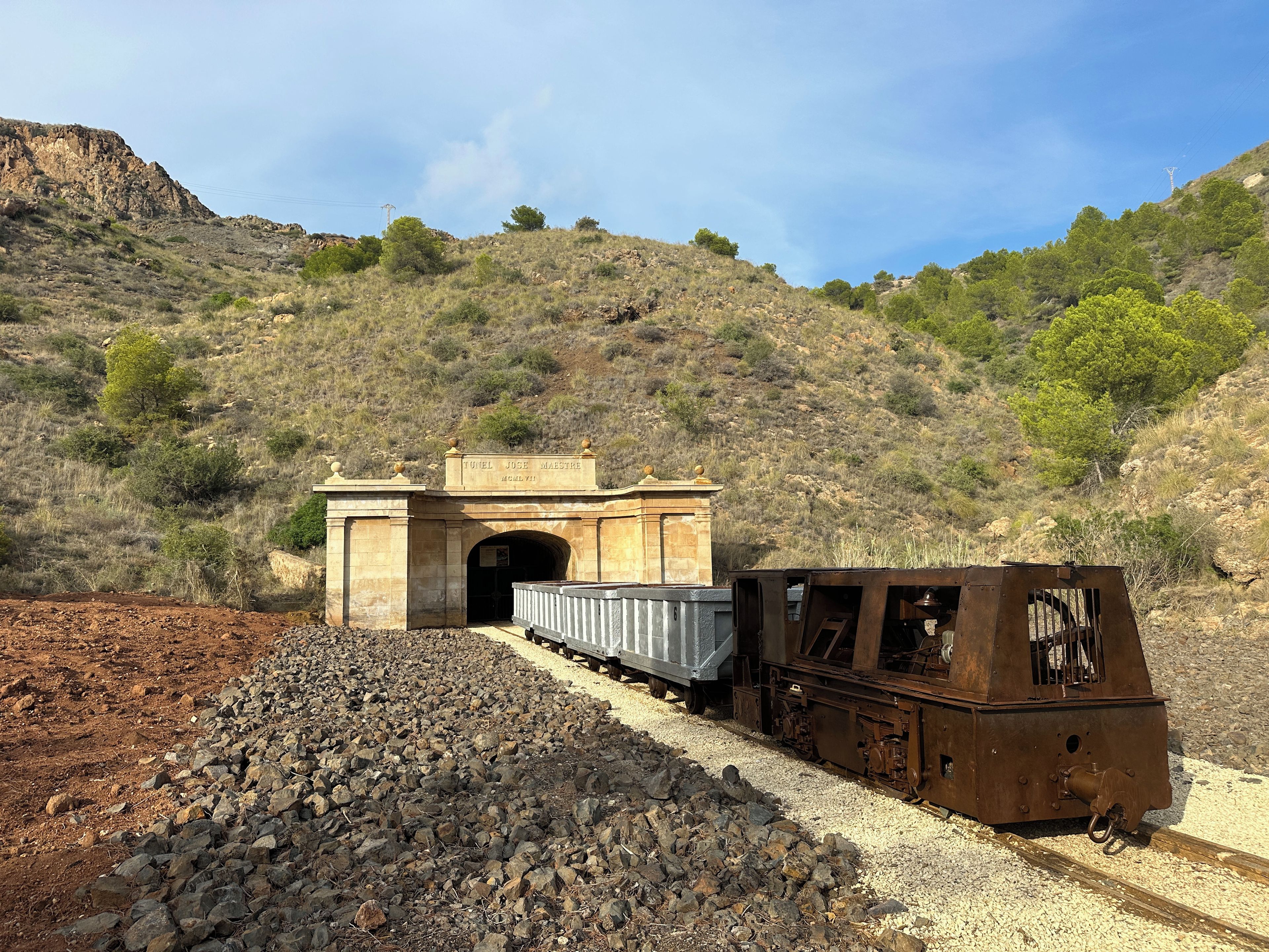 Estação de trem com entrada de túnel, caminho de cascalho e um morro rochoso ao fundo.