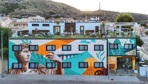 A mural with a classical figure, a modern building with a flat roof, and a hotel with a visible entrance. - Hotel Portmán (La Union)