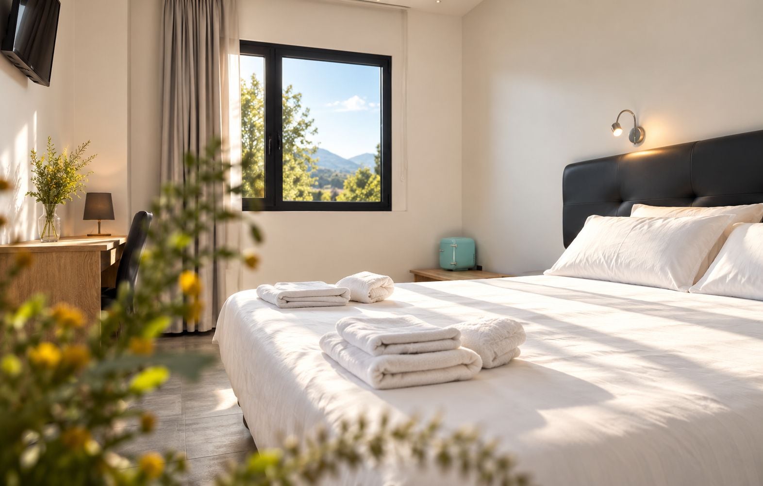 Una camera d'hotel con un letto, una scrivania con una lampada, una finestra vista montagne e un comodino con una lampada.