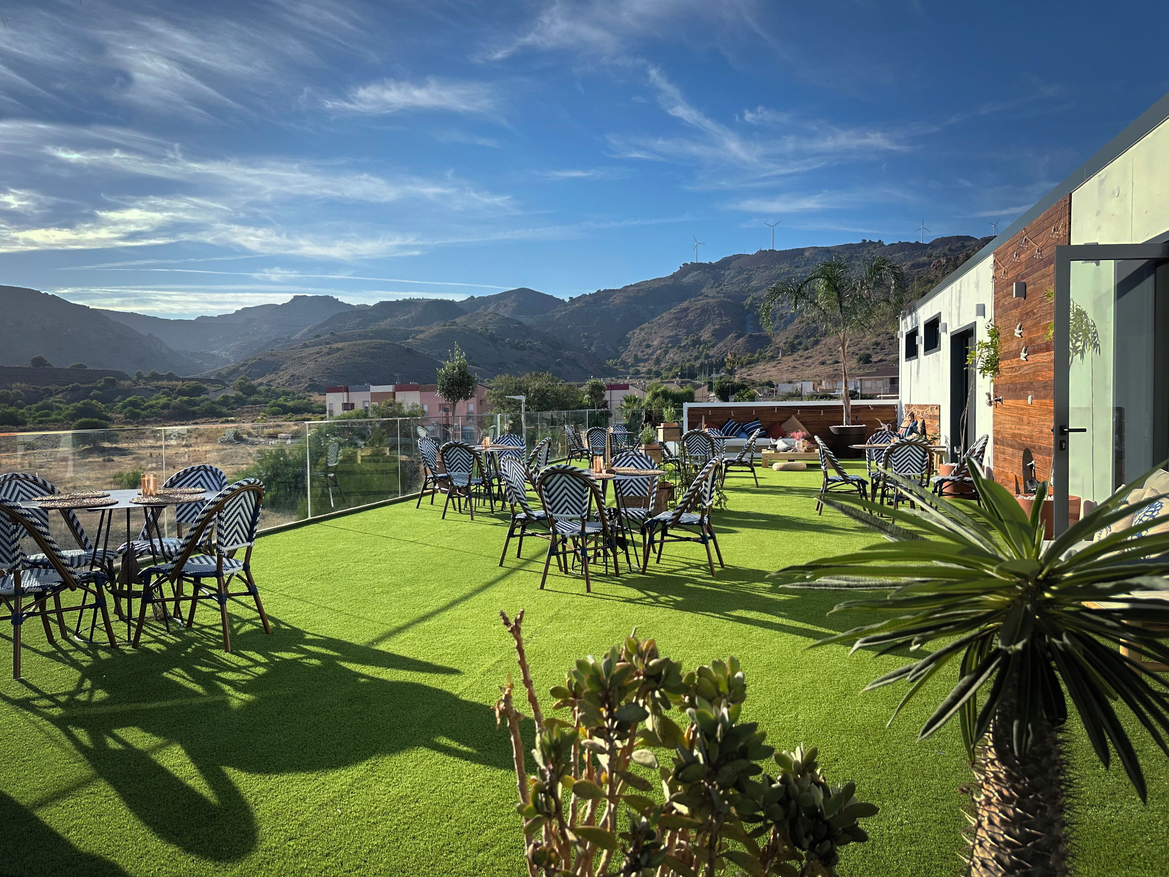 An outdoor seating area with chairs and tables, a building with a wooden facade, and a view of mountains in the background.