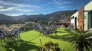 An outdoor seating area with chairs and tables, a building with a wooden facade, and a view of mountains in the background.