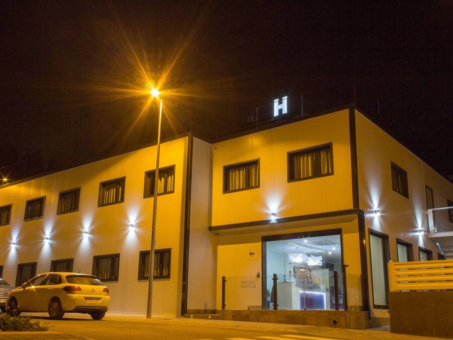 Un edificio moderno con un llamativo letrero en forma de 'H', que indica un hospital. Cuenta con múltiples ventanas, una entrada de vidrio y un área de estacionamiento con automóviles.