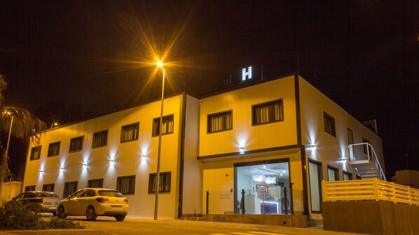 Un edificio moderno con un llamativo letrero en forma de 'H', que indica un hospital. Cuenta con múltiples ventanas, una entrada de vidrio y un área de estacionamiento con automóviles.