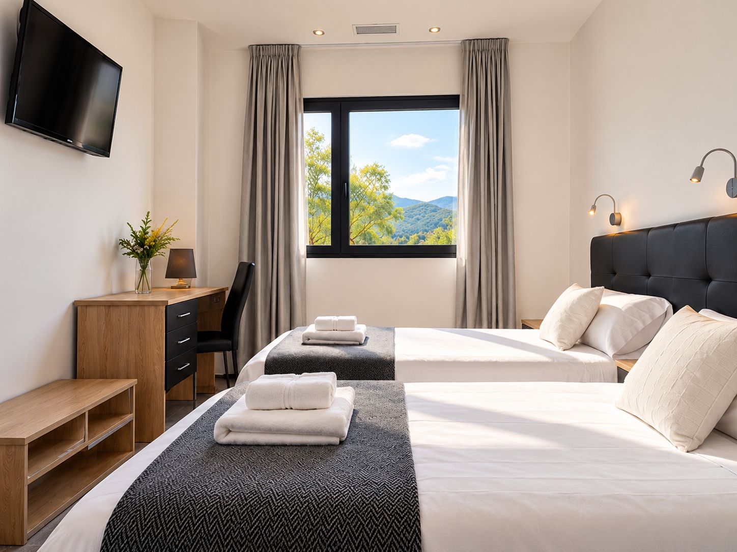 Habitación de hotel con dos camas, un escritorio, una silla, un televisor y una ventana con vistas a árboles y montañas.