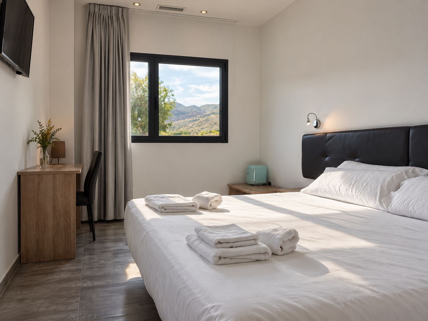 Habitación de hotel moderna con una cama grande, un escritorio con lámpara, vista al exterior y una maleta en la mesita de noche.