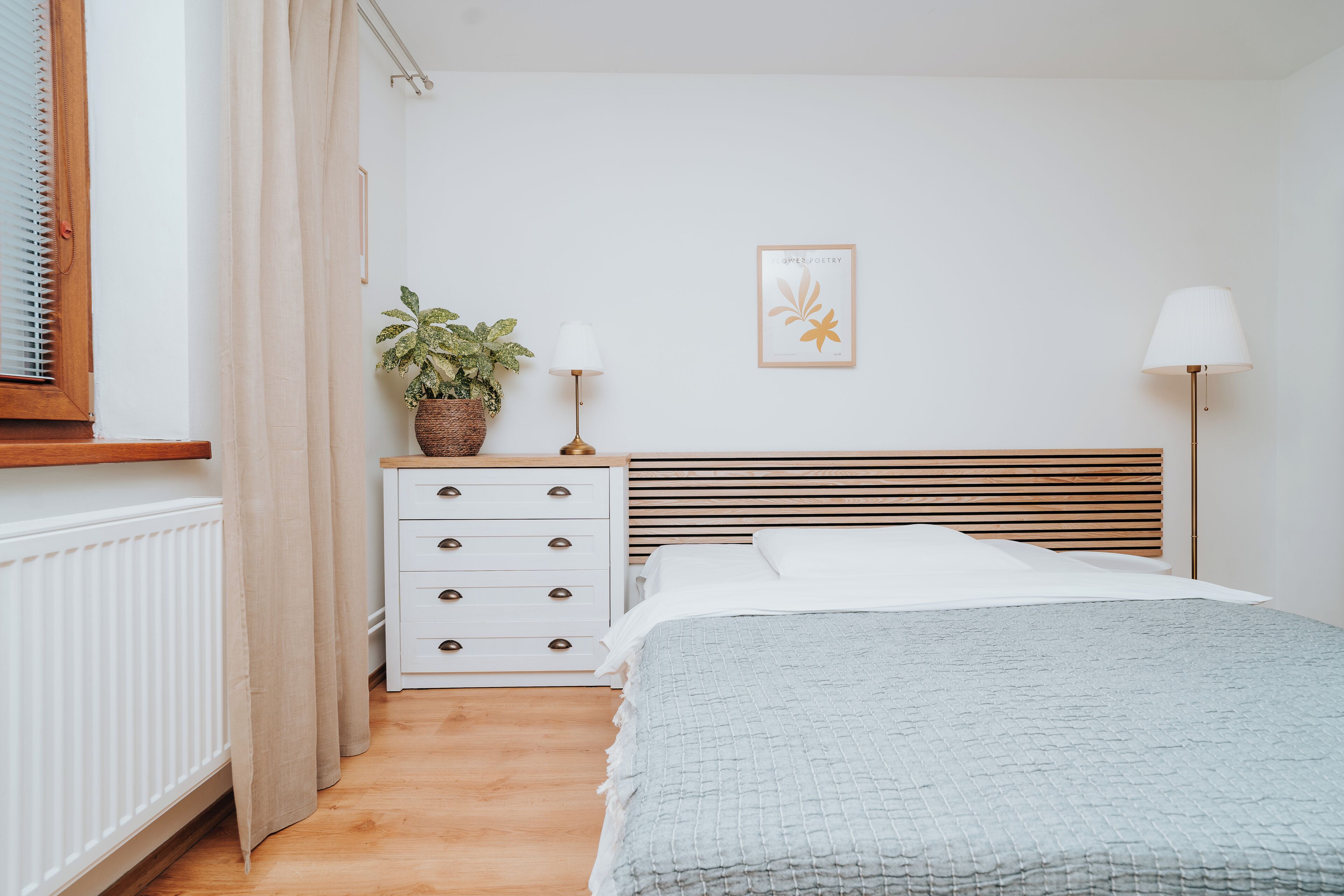 A bedroom with a wooden headboard, a white bedside table with drawers, a potted plant, a framed picture on the wall, and a bedside lamp.