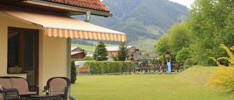 A house with a red-tiled roof, a covered patio with wicker furniture, and a view of a green landscape with a playground and mountains in the background.