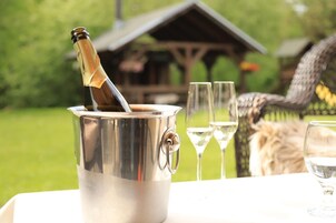 Un balde metálico con una botella de champán, dos copas de vino vacías y una silla de mimbre en un jardín.