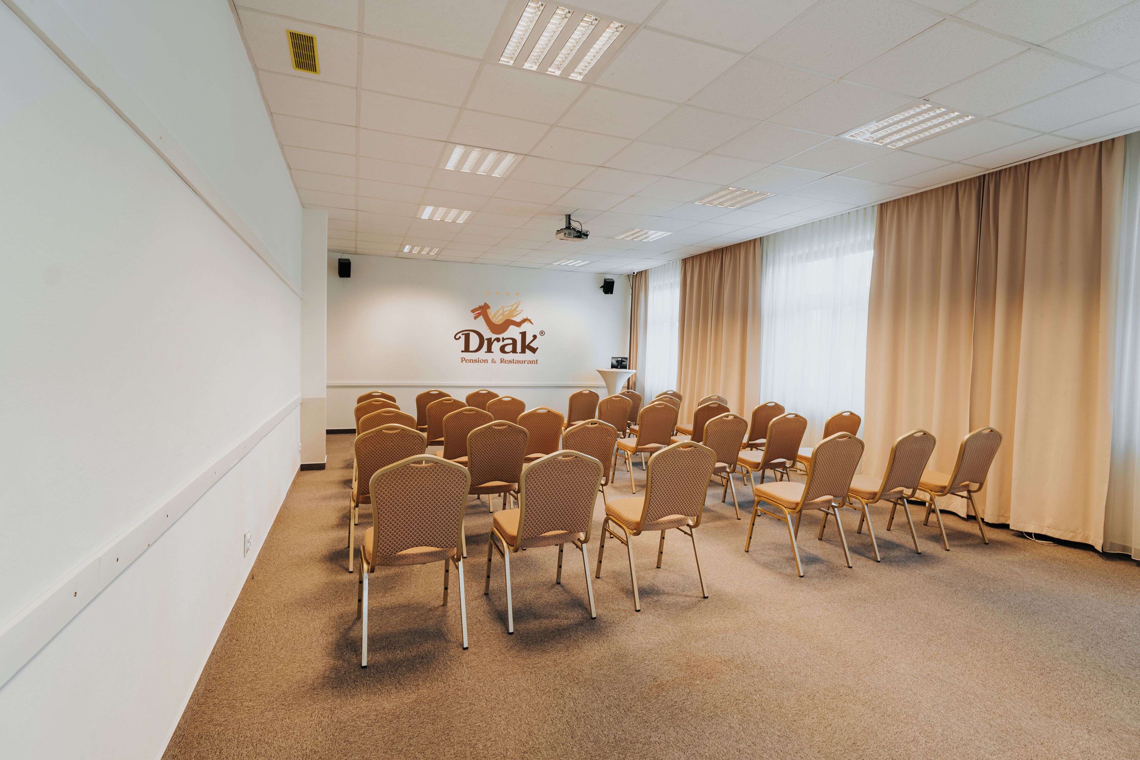 A conference room with rows of chairs, a whiteboard with the logo 'Drak Pension & Restaurant', and a projector on the ceiling.
